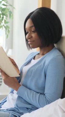Yatakta oturmuş kitap okuyan odaklanmış Afro-Amerikan bir kadının portresi. Evde bir hikayenin tadını çıkarırken meraklı ve düşünceli görünüyor..