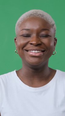 Kısa sarı saçlı, gülen ve gözleri kapalı gülümseyen neşeli Afro-Amerikan kadın. Yeşil arkaplanda dikey video izole edildi.