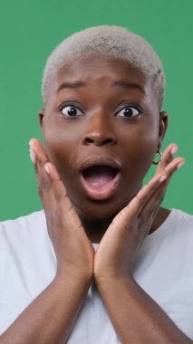 Genç Afro-Amerikalı kadın şaşırmış görünüyor. Elleri yüzünde, sonra gülümsüyor. Yeşil arkaplanda dikey video izole edildi.