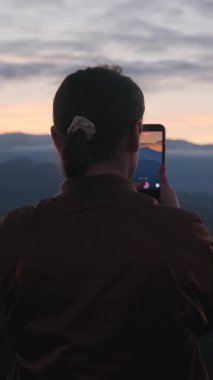 İspanya 'nın Asturias şehrinde gün batımının fotoğraflarını çeken bir kadının arka görüntüsü. Manzarayı yakalayan dikey video.