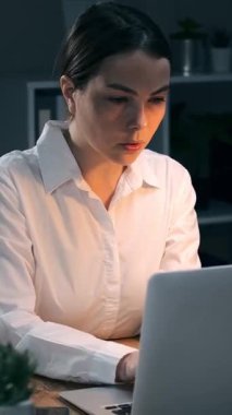 Gece geç saatlerde karanlık ofiste dizüstü bilgisayarla çalışan genç bir iş kadını. Çok çalışan kadın daktilo. Dikey video.