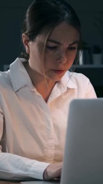 Stresli genç iş kadını gecenin geç saatlerinde karanlık ofiste dizüstü bilgisayarla sorun yaşıyor. Çok çalışan bayan çalışanlar yorgun. Dikey video.