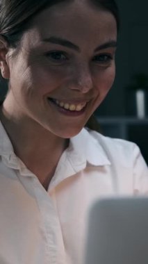 Gecenin geç saatlerinde karanlık ofiste dizüstü bilgisayar ekranına bakan gülümseyen genç iş kadınının yakın çekimi. Mutlu bayan çalışan işten zevk alıyor. Dikey video.