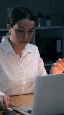 Gece geç saatlerde karanlık bir ofiste dizüstü bilgisayar başında çalışırken elinde kalem tutan düşünceli genç bir iş kadını. Çok çalışan kadın çalışan. Dikey video.