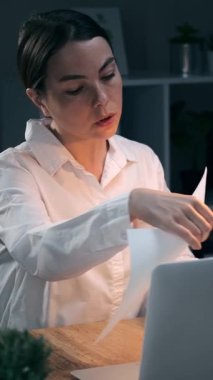 Gece geç saatlere kadar karanlık ofiste çalışan, kağıt belgelerini kontrol eden ve dizüstü bilgisayarda yazan genç bir iş kadını. Çok çalışan kadın çalışan. Dikey video.