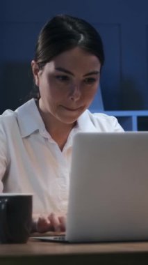 Gülümseyen genç iş kadını kahve içiyor ve gece geç saatlerde karanlık ofiste dizüstü bilgisayarda çalışıyor. Mutlu kadın çalışan. Dikey video.