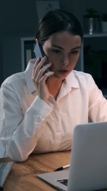 Gülümseyen genç iş kadını cep telefonuyla konuşuyor ve gecenin geç saatlerinde karanlık ofiste dizüstü bilgisayarla çalışıyor. Bayan bir çalışanımız arıyor. Dikey video.
