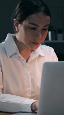 Gece geç saatlere kadar karanlık ofiste çalışan heyecanlı genç iş kadını başarıyı ya da iyi haberi laptopta kutluyor. Mutlu bayan çalışan kazanıyor. Dikey video.