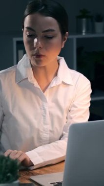 Gece geç saatlere kadar karanlık ofiste çalışan, kağıt belgelerini kontrol eden ve dizüstü bilgisayarda yazan genç bir iş kadını. Çok çalışan kadın çalışan. Dikey video.
