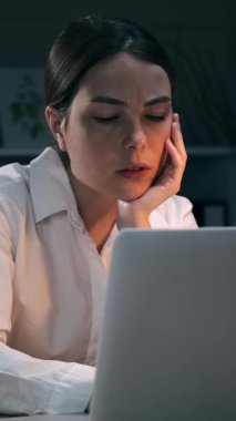 Yorgun genç iş kadını gece geç saatlerde dizüstü bilgisayarda çalışırken gözlerini ovuşturuyor. Çok çalışan bayan çalışanın uykusu geliyor. Dikey video.