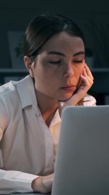 Yorgun genç iş kadını kollarını açıyor, uyanıyor ve gece geç saatlerde dizüstü bilgisayarında çalışmaya devam ediyor. Çok çalışan bayan çalışanın enerjisi geri geliyor. Dikey video.