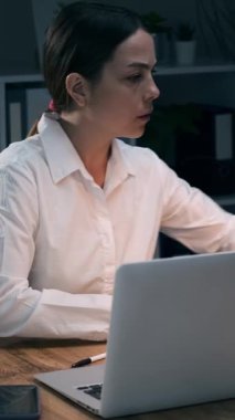 Gece geç saatlere kadar karanlık ofiste çalışan, dizüstü bilgisayarda yazan ve cep telefonuyla konuşan genç bir iş kadını. Çok çalışan kadın çalışan. Dikey video.