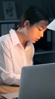 Gece geç saatlere kadar karanlık ofiste çalışan, kalem ve dizüstü bilgisayarla kağıt dökümanlarını kontrol eden genç bir iş kadını. Çok çalışan kadın çalışan. Dikey video.