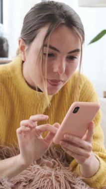 Genç bir kadın akıllı telefondan mesaj okuyor ve evdeki kanepede yatarken birden kafası karışmış ya da endişeli görünüyor. Dikey video.