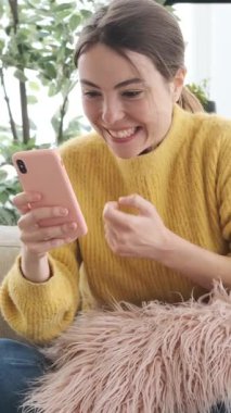 Heyecanlı genç bir kadın akıllı telefonu kontrol ediyor, zaferde yumruğunu sıkıyor ve koltukta otururken başarıyı veya iyi haberi kutluyor. Dikey video.