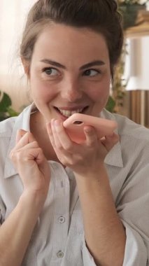 Neşeli genç bir kadın akıllı telefondan sesli bir mesaj kaydediyor. Gülümsüyor ve telefonu ağzının yanında tutarken heyecanla el sallıyor. Dikey video.
