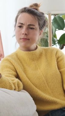 Akıllı telefondan konuşan, gülen, çay içen ve kanepede otururken saçını düzelten neşeli genç bir kadın. Dikey video.