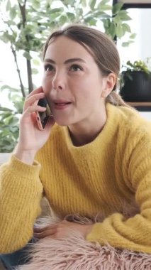 Akıllı telefondan konuşan neşeli genç bir kadın sohbet sırasında gülüyor ve iyi haberi kanepede yumruk hareketiyle kutluyor. Dikey video.