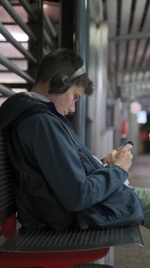 Kulaklık takmış, gece vakti tren istasyonundaki bir bankta oturup akıllı telefonuna bakan yorgun bir gencin dikey videosu..