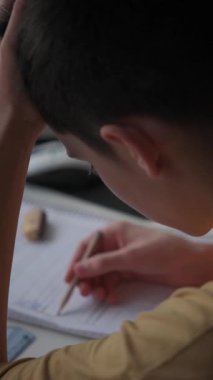 Bir gencin kalemle not defterine yazıp evde ders çalışırken zor bir problemi düşünmesini gösteren dikey bir video..