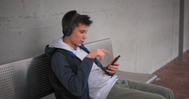 Neşeli genç bir çocuk metro bankında oturmuş, kulaklık takmış bir arkadaşına telefon görüşmesini yaparken gülüyor ve ekranı işaret ediyor..