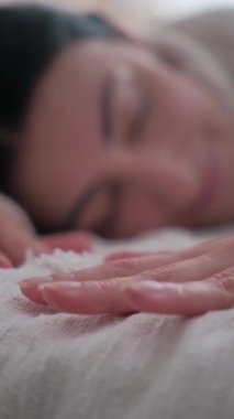 Kadın elinin yatakta yumuşak beyaz battaniye dokusuna nazikçe dokunması. Bulanık gülümseyen kadın uyuyor ya da arka planda uyanıyor. Dikey video.