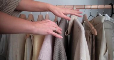 Asılı giysileri sınıflandıran ve askıdan bej süveter seçen kadın ellerini kapat. Kadın yatak odasında kıyafet seçiyor..