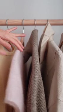 Asılı giysileri sınıflandıran ve askıdan bej süveter seçen kadın ellerini kapat. Yatak odasında kıyafet seçen bir kadın. Dikey video.