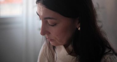 Pencerenin yanındaki cam bardaktan sıcak kahve içen düşünceli, koyu renk saçlı kadını yakından çek. Kadın kahvesini yudumluyor ve evde dalgın dalgın bakıyor..