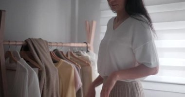 Koyu renk saçlı kadın elbise rafından beyaz yün süveteri alıp üzerine giyiyor. Kadın, evdeki parlak yatak odasında hazırlanıyor..