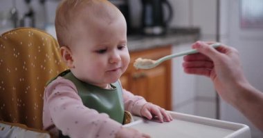 Neşeli bir kız çocuğu yüksek bir sandalyede oturur, annesinin kaşığından yer ve ellerini mutlu bir şekilde çırpar. Sağlıklı beslenme ve erken gelişim kavramı.