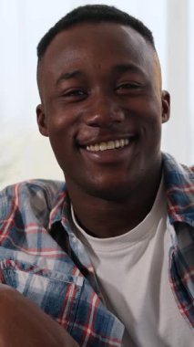 Dikey video. Genç, Afro-Amerikan bir adamın gülüşünün portresi. Kameraya neşeli bir ifadeyle bakıyor. Parlak bir iç mekanda..