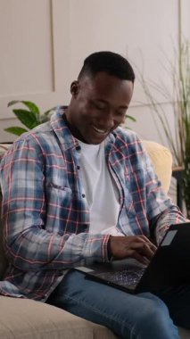 Neşeli bir siyahi adam oturma odasındaki bir kanepede dinlenirken dizüstü bilgisayarda gülümseyip yazı yazar. Dikey video.