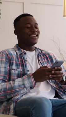 Genç ve mutlu bir Afro-Amerikalı adamın cep telefonuyla mesajlaştığı ve evdeki kanepede otururken güldüğü dikey bir video..