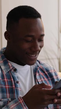 Genç, mutlu, Afro-Amerikan bir adamın evde akıllı telefonunu kullanırken güldüğü ve şaşırdığı dikey bir video..
