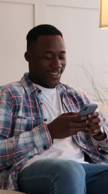 Evdeki bir kanepede akıllı telefonunu kullanırken gülen neşeli bir gencin dikey videosu. İnternetteki komik içerikten hoşlanıyor..