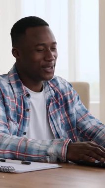 Mutlu Afro-Amerikalı bir adamın dizüstü bilgisayarında bir video araması var. Evde bir masada otururken gülümsüyor ve el kol hareketi yapıyor. Dikey video.