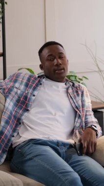 Kanepede akıllı telefonunu kullanan sıkılmış bir Afrikalı Amerikalı 'nın dikey videosu. Beklerken hayal kırıklığına uğramış görünüyor ve sonra kanepeye uzanıyor..