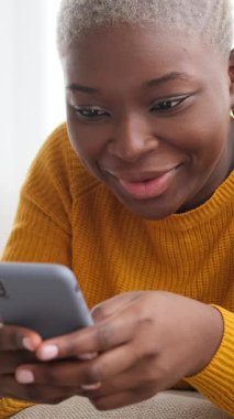 Mutlu Afrikalı Amerikalı kadın cep telefonunu evde kullanıyor. O, sosyal medyada mesajlaşırken veya gezinirken gülümsüyor. Dikey video.