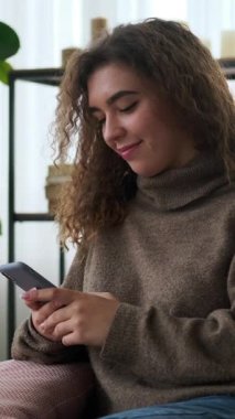Evde akıllı telefonuyla mesajlaşırken gülümseyen ve gülen kıvırcık saçlı mutlu genç bir kadının dikey videosu..