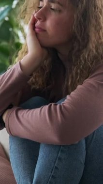 Üzgün ve depresif genç bir kadının evde yalnız başına oturup umutsuz, yalnız ve endişeli hissetmesinin dikey videosu..