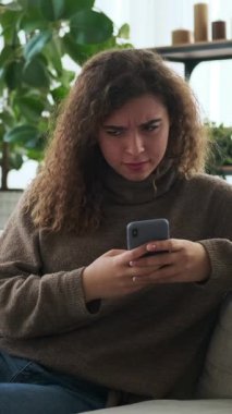 Bir kadının şaşkınlıkla tepki verdiği ve evdeki koltukta akıllı telefonunu kullanırken gülümsediği dikey bir video. Sosyal medyada mesajlaşıyor ve geziniyor..