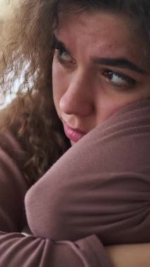 Kıvırcık saçlı üzgün ve düşünceli genç bir kadının dikey videosu. Duygusal yüzüne yakın çekim, melankoli ve depresyon duygularını gösteriyor..