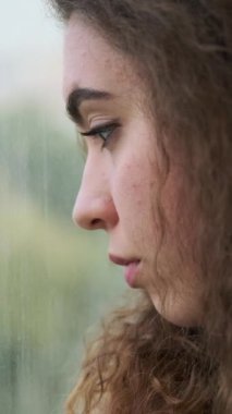 Camdan dışarı bakan, üzgün ve yalnız hisseden düşünceli genç bir kadının yakın plan profili. Dikey video.