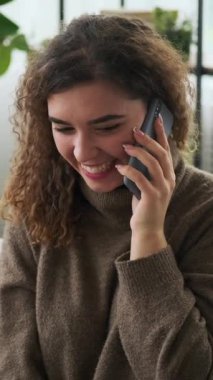 Evde cep telefonuyla konuşurken gülümseyen ve gülen kıvırcık saçlı mutlu genç bir kadın. Dikey video.
