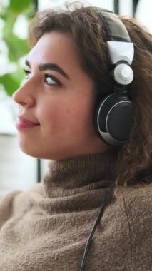 Kulaklık takan mutlu genç bir kadın en sevdiği müziği dinliyor, evde dinleniyor. Dikey video.