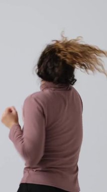 Kıvırcık saçlı mutlu genç bir kadın enerjik bir şekilde dans eder, beyaz bir arka planda izole bir şekilde. Dikey video.