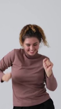 Kıvırcık saçlı, enerjik dans eden, gülümseyen, beyaz arka planda soyutlanmış mutlu bir genç kadın. Dikey video.