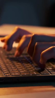 Karanlık bir odada, dizüstü bilgisayarda sıcak ışıkla yazı yazan ellerin yakın çekimi. Dikey video.