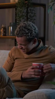 Evdeki kanepede oturup film izlerken gülen ve kahve içen genç bir adamın dikey videosu..